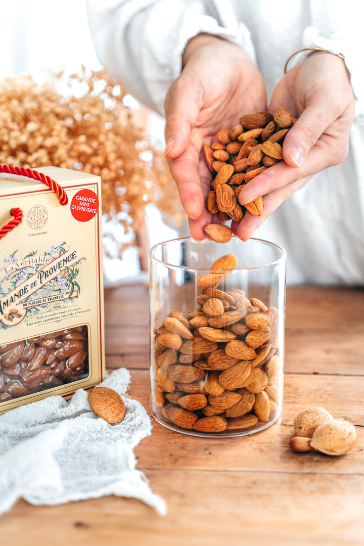 L'image montre une personne versant des amandes entières dans un bocal en verre. Les amandes sont de couleur brun doré, et l'on voit également quelques coquilles d'amandes autour du bocal. À gauche de l'image, une boîte d'amandes de Provence de la marque Terraroma est visible, avec une étiquette "Garantie sans glyphosate". L'ambiance est chaleureuse, avec un arrière-plan flou qui semble contenir des fleurs séchées et des éléments en bois, renforçant l'atmosphère artisanale et naturelle.