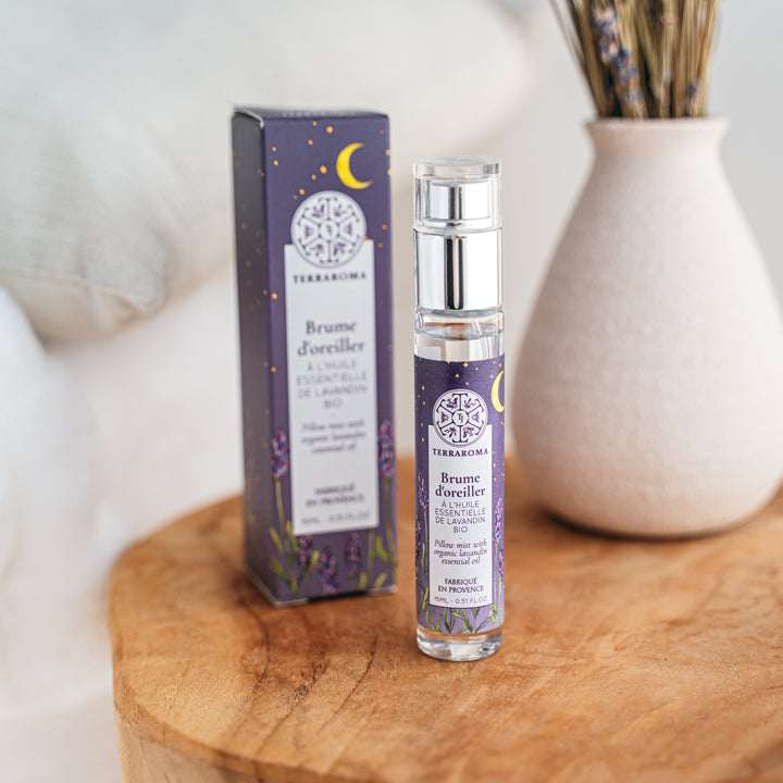 La photo montre un flacon de brume d'oreiller à l'huile essentielle de lavande bio de 15 ml, posé sur une table en bois clair. Le flacon est élégant, transparent, et son étiquette violette est décorée de motifs de lavande et d'une lune dorée. À l'arrière, on distingue la boîte assortie, ainsi qu'un vase blanc contenant des brins de lavande séchés, créant une ambiance douce et apaisante.