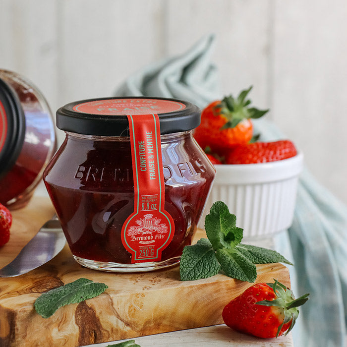 Un pot en verre de confiture de fraise et menthe de la marque Bremond Fils, avec un couvercle noir et une étiquette rouge indiquant 'Confiture fraise & menthe' et un poids net de 250 g. Le pot est posé sur une planche en bois avec quelques fraises fraîches et des feuilles de menthe autour. À l'arrière-plan, il y a un petit bol contenant des fraises, ajoutant une touche de fraîcheur à la présentation.