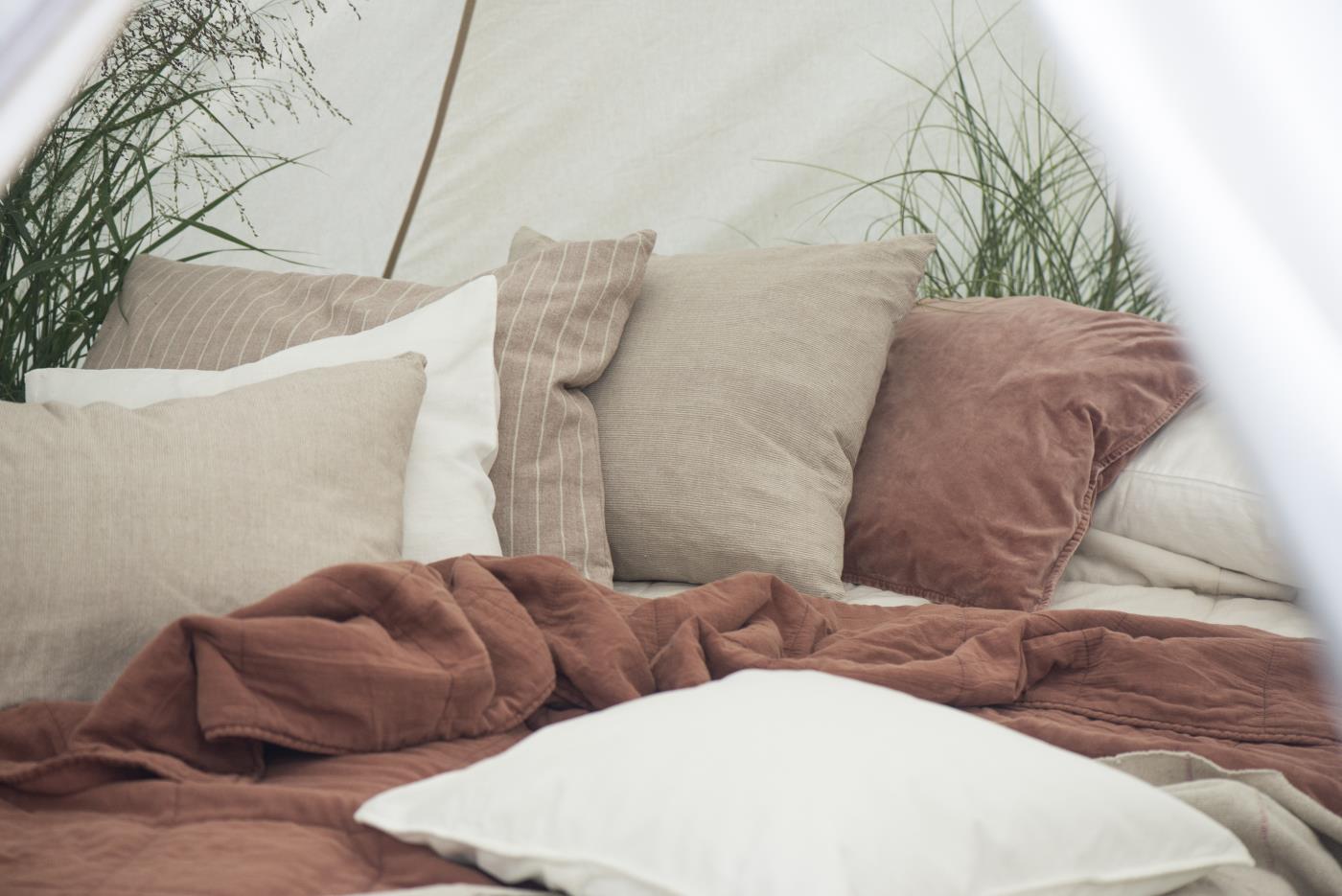 Une scène paisible montrant un lit avec plusieurs coussins dans des tons naturels. Les coussins sont disposés sur des draps en lin beige et rouille, créant une ambiance douce et chaleureuse. En arrière-plan, des plantes vertes ajoutent une touche naturelle et apaisante à l’ensemble.