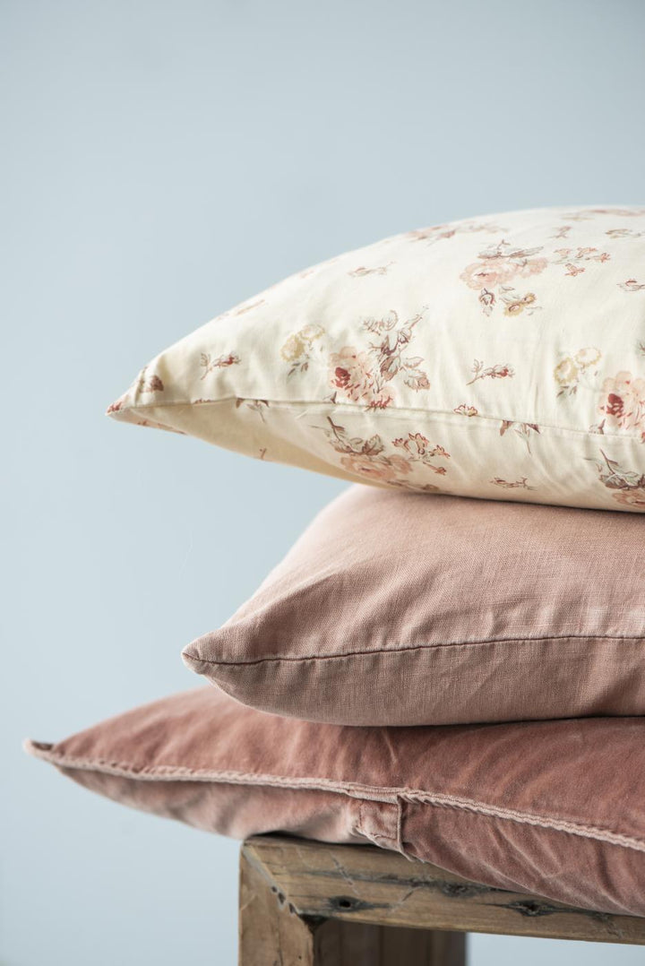 Trois coussins empilés sur une surface en bois rustique. Le coussin du bas est en velours couleur rouille, celui du milieu en lin rose, et celui du dessus présente un motif floral sur un fond crème, offrant un mélange de textures douces et naturelles.