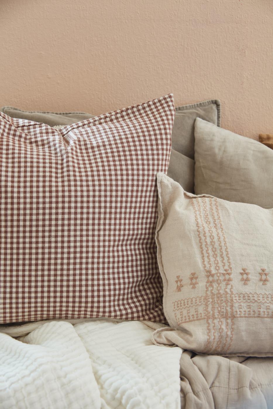 Cette image montre deux coussins disposés sur un lit ou un canapé, créant une ambiance cosy et chaleureuse. Le premier coussin est à carreaux marron et blanc, avec un motif vichy classique, qui ajoute une touche rustique et rétro. Le deuxième coussin est plus neutre, avec un motif brodé discret dans des tons beige et crème, offrant un contraste doux et élégant avec le premier coussin. Cette combinaison de textures et de motifs apporte une atmosphère accueillante et confortable.