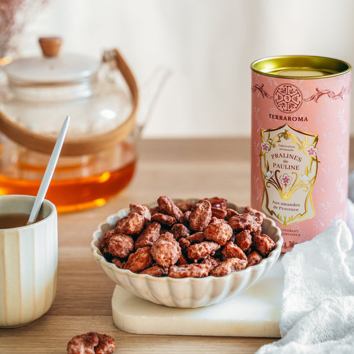 L'image montre un bol rempli de pralines aux amandes de Provence, placées devant un contenant rose de la marque Terraroma avec l'inscription "Pralines de Pauline". Le récipient est élégant, décoré de motifs floraux, et met en avant la fabrication artisanale du produit. À gauche du bol, on aperçoit une tasse de thé et une théière en verre contenant une boisson chaude couleur ambrée, créant une ambiance de dégustation chaleureuse et réconfortante. 