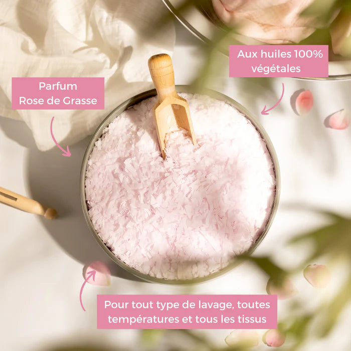 Un bol contenant des flocons de savon rose de Grasse. Le bol est en métal et rempli de flocons de savon rose clair, accompagnés d'une petite pelle en bois. Des éléments textuels autour du bol indiquent que le produit est parfumé à la rose de Grasse, qu'il est composé d'huiles 100% végétales et qu'il convient pour tout type de lavage, à toutes les températures et pour tous les tissus. En arrière-plan, des éléments naturels, tels que des pétales de rose et des tissus en lin.