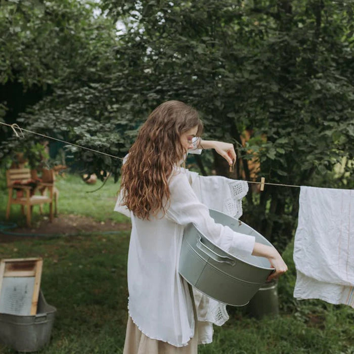 Une femme aux cheveux longs et ondulés étend du linge à l'extérieur sur une corde à linge dans un jardin verdoyant. Elle porte une blouse blanche fluide et tient une grande bassine en métal. À l'arrière-plan, on aperçoit des meubles en bois sous des arbres, créant une atmosphère paisible et naturelle.