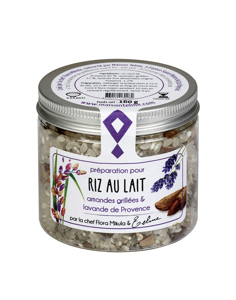 Le pot en verre transparent contient une préparation pour riz au lait, agrémentée d'amandes grillées et de lavande de Provence. L'étiquette est ornée de dessins de brins de lavande violet et d'amandes, évoquant les ingrédients principaux de cette recette provençale. Sur le couvercle métallique argenté, les instructions de préparation sont indiquées en détail, ainsi que le poids net de 160 grammes. La mention "par la chef Flora Mikula & Telme" met en avant l'authenticité artisanale et l'expertise culinaire.