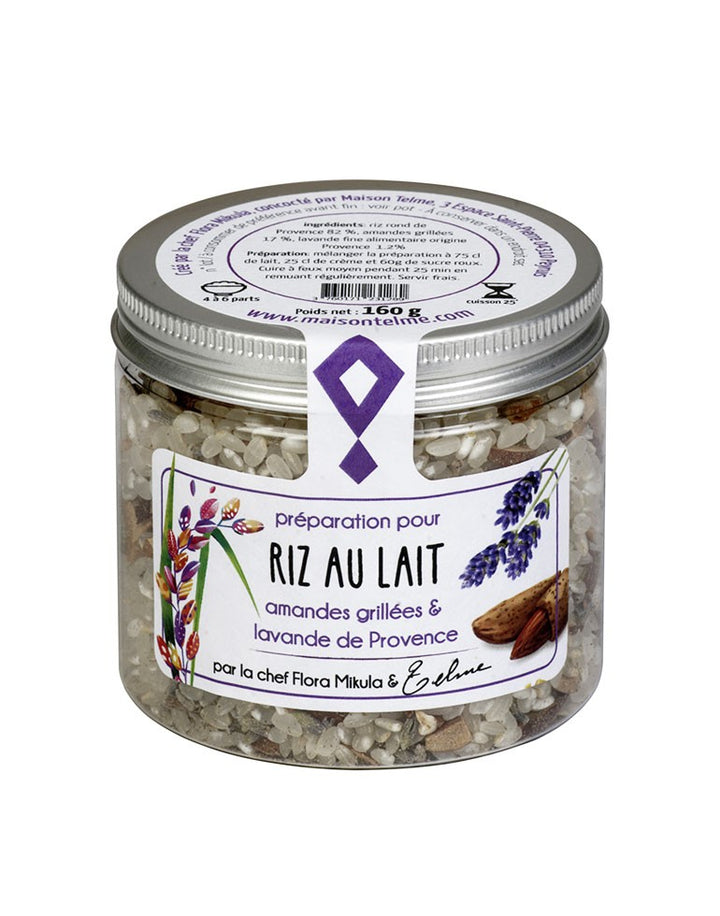 Le pot en verre transparent contient une préparation pour riz au lait, agrémentée d'amandes grillées et de lavande de Provence. L'étiquette est ornée de dessins de brins de lavande violet et d'amandes, évoquant les ingrédients principaux de cette recette provençale. Sur le couvercle métallique argenté, les instructions de préparation sont indiquées en détail, ainsi que le poids net de 160 grammes. La mention "par la chef Flora Mikula & Telme" met en avant l'authenticité artisanale et l'expertise culinaire.