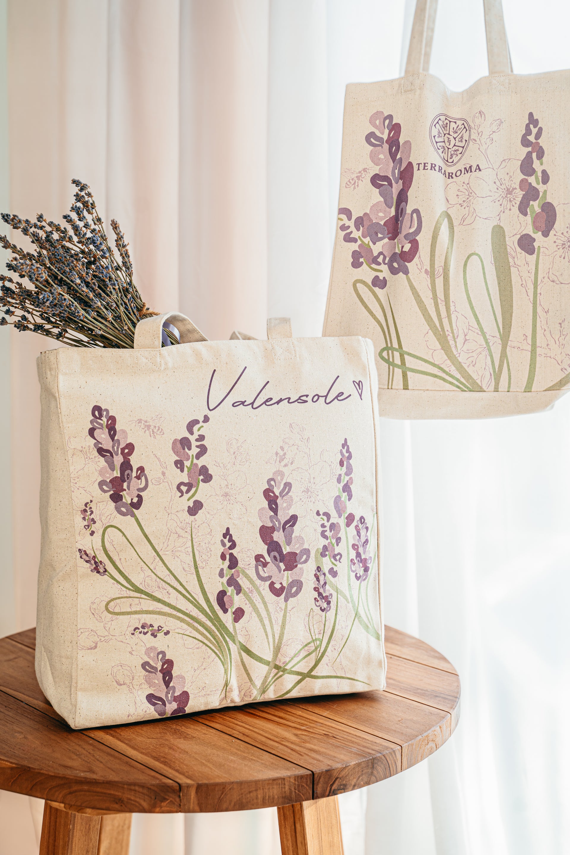 L'image montre deux sacs en tissu décorés de motifs de lavande, associés à la marque Terraroma. Le sac au premier plan contient un bouquet de lavande et arbore l'inscription manuscrite « Valensole » avec un petit cœur à côté, ainsi qu'un motif de lavande violette accompagné de feuilles vertes. L'arrière-plan présente un second sac suspendu, où l'on distingue également un motif de lavande avec le logo de Terraroma visible en haut. Les sacs sont posés ou accrochés dans un environnement lumineux.