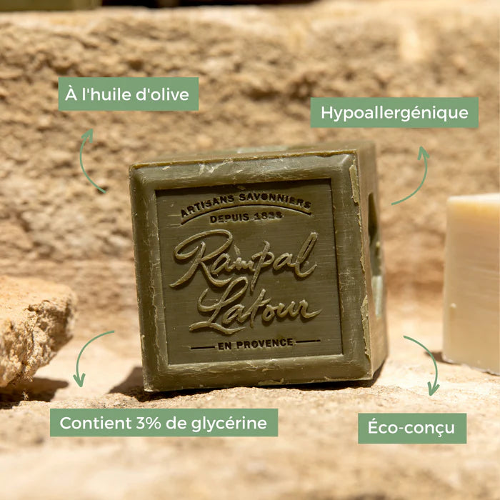 Un savon artisanal de la marque Rampal Latour, fabriqué à l'huile d'olive. Le savon est de couleur verte et se présente sous la forme d'un cube. Des annotations indiquent que le savon est hypoallergénique, qu'il contient 3 % de glycérine et qu'il est éco-conçu. Le fond de l'image montre une texture naturelle, probablement en pierre, qui met en valeur le produit.