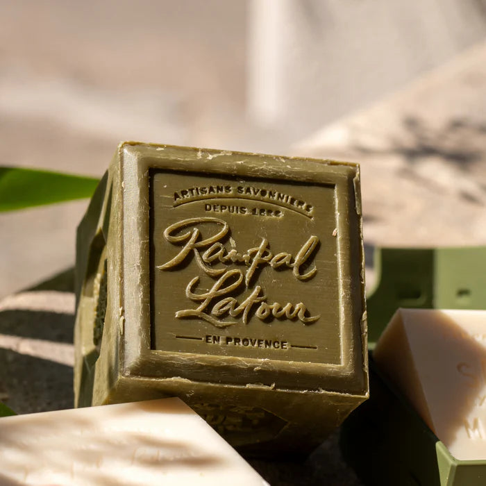 Un savon artisanal de la marque Rampal Latour, fabriqué à l'huile d'olive. Le savon est présenté sous la forme d'un cube vert, avec le logo de la marque et les inscriptions 'Artisans Savonniers depuis 1828' et 'en Provence'. Le savon est posé sur une surface en pierre, entouré de quelques feuilles vertes et d'autres morceaux de savon de couleur claire.