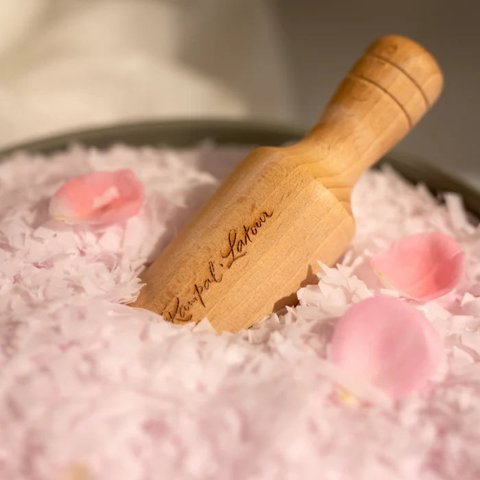 Un gros plan d'une petite pelle en bois gravée avec le nom 'Rampal Latour', posée sur un lit de flocons de savon roses. Des pétales de rose se trouvent également dispersés parmi les flocons, ajoutant une touche décorative à la composition.