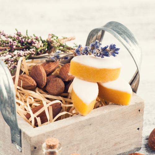 L'image présente une petite caisse en bois rustique remplie de calissons, d'amandes entières et de brins de lavande séchée. Les calissons, alignés au sommet, se distinguent par leur glaçage blanc et leur base dorée. Le tout est niché sur un lit de raphia, dans une ambiance douce et naturelle évoquant la Provence.