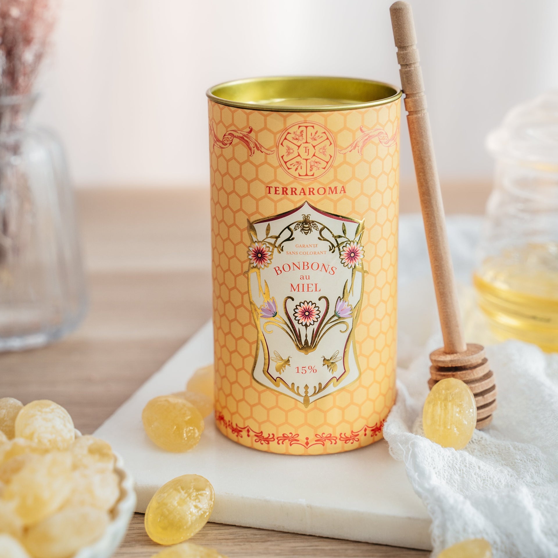 L'image présente une boîte cylindrique jaune de la marque Terraroma contenant des bonbons au miel (15 %). La boîte est décorée d'un motif en nid d'abeilles et d'une étiquette florale raffinée. Autour, des bonbons dorés, une cuillère en bois pour le miel, et un pot de miel ajoutent une touche naturelle et chaleureuse, le tout dans une ambiance lumineuse et épurée.