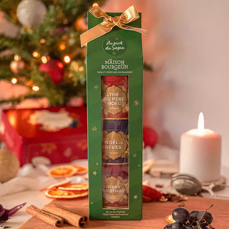 Coffret cadeau "Au pied du Sapin" de la Maison Bourgeon, présenté dans une ambiance festive avec un ruban doré. Le coffret contient trois thés de Noël : "Le Thé du Père Noël", "Noël en Rêves" et "Cherry Christmas". Le paquet vert décoré de flocons de neige est mis en scène devant un sapin illuminé, entouré d'éléments chaleureux comme une bougie, des bâtons de cannelle, et des tranches d'orange séchées, évoquant l'atmosphère magique et réconfortante des fêtes de fin d'année.