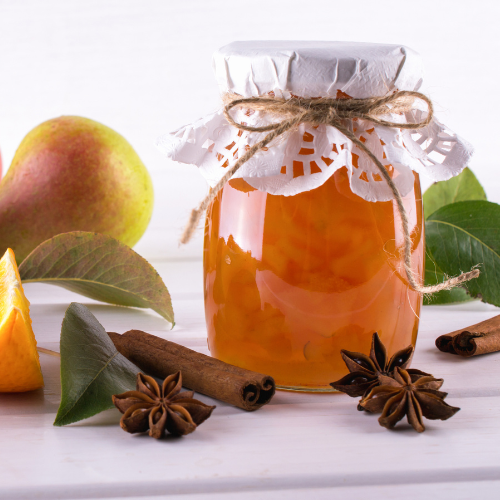 L'image montre un pot de confiture maison au look artisanal, décoré d'un couvercle en papier dentelé attaché avec une ficelle. Autour du pot, des bâtons de cannelle, des étoiles de badiane, des feuilles, des poires et des morceaux d'orange évoquent les ingrédients ou accompagnements possibles. La scène est lumineuse, avec un fond blanc qui met en valeur les teintes chaudes de la confiture.