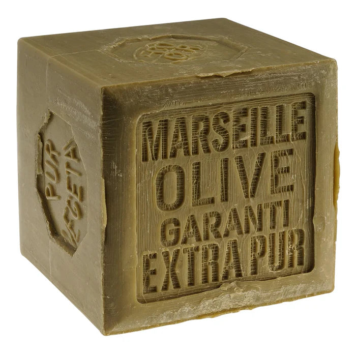 Un savon en cube de la marque Rampal Latour, étiqueté comme 'Marseille Olive Garanti Extrapur'. Le savon a une couleur vert olive et présente une texture légèrement rugueuse. Les inscriptions sur le savon indiquent qu'il est fait à partir d'huile d'olive et garantit sa pureté. Le savon est posé sur une surface neutre, mettant en valeur son aspect artisanal.