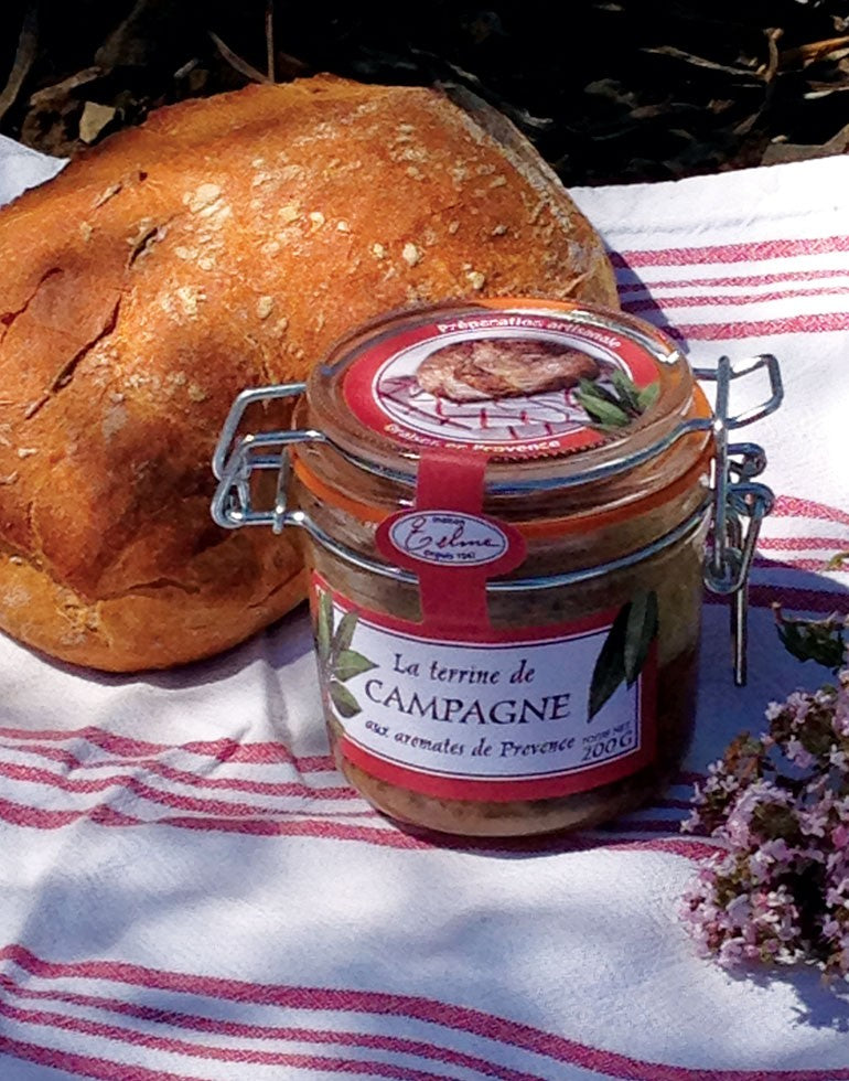 Terrine de Campagne aux Aromates de Provence de la Maison Telme, posée sur une nappe à rayures rouges et blanches, à côté d'une miche de pain rustique. Le cadre évoque un pique-nique en pleine nature, avec des touches de thym fleuri, mettant en valeur le caractère artisanal et les saveurs provençales de cette spécialité.