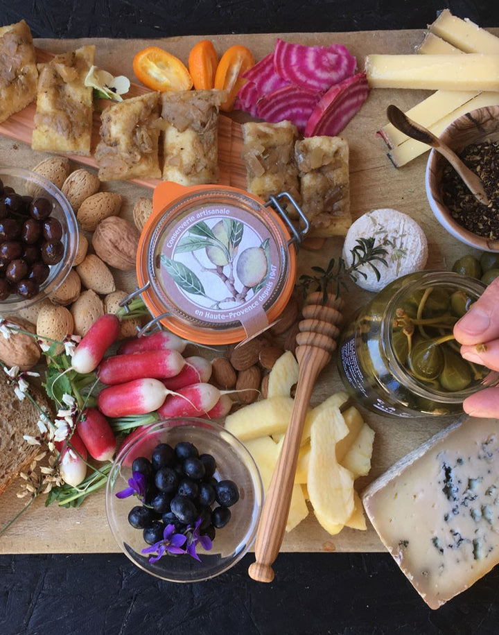 Planche apéritive gourmande comprenant la Terrine aux Amandes de Haute-Provence de la Maison Telme. La terrine est entourée d'une variété de délices : radis, myrtilles décorées de fleurs violettes, tranches de pommes, morceaux de fromage, noix, amandes, olives, morceaux de pain, et tranches de betterave chioggia. Cette présentation met en avant la richesse et la diversité des produits provençaux pour un moment de partage convivial.