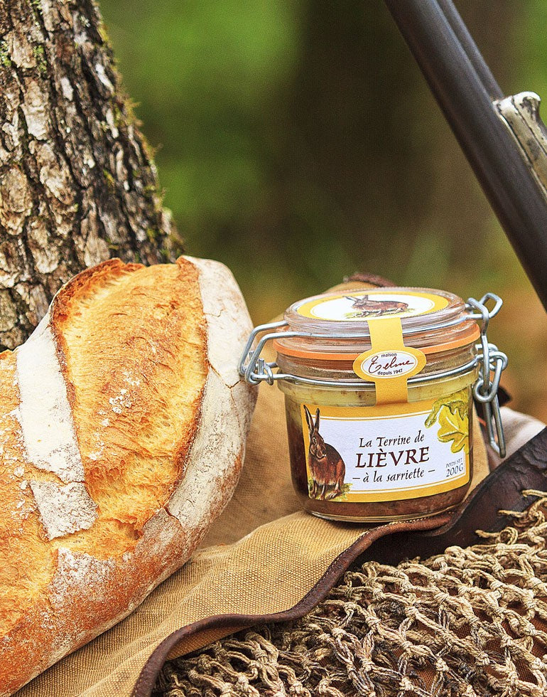 Terrine de Lièvre à la Sarriette de la Maison Telme, présentée dans un cadre champêtre. Le pot en verre est accompagné d'une baguette rustique, posé sur un tissu et un filet, évoquant un repas en pleine nature. L'étiquette jaune arbore une illustration de lièvre, renforçant le caractère authentique et régional de cette spécialité artisanale de Haute-Provence.